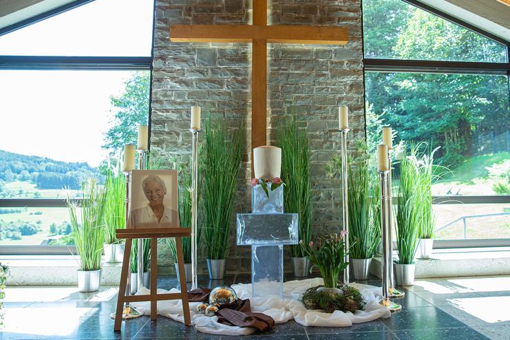 Ein Holzkreuz, eine blühende Pflanze und ein Foto einer älteren Frau in einer lichtdurchfluteten Kapelle.