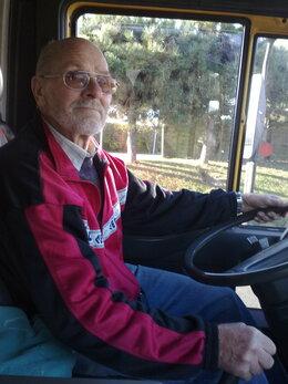 Ein älterer Mann mit Brille sitzt am Steuer eines Fahrzeugs und blickt in die Kamera, im Hintergrund Bäume sichtbar.