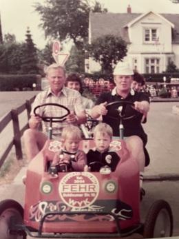 Familie fährt in einem roten Pedalauto, zwei Kinder vorne, Erwachsene hinten, im Hintergrund ein Haus und Verkehrszeichen.