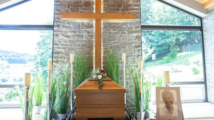 Elegante Trauerfeier mit einem geschmückten Holzsarg, Kerzen und einem Kreuz in einem hellen Raum mit Aussicht.