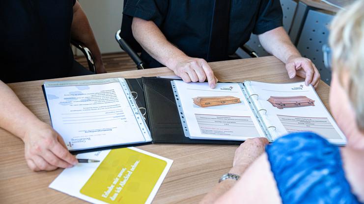 Drei Personen am Tisch betrachten Katalogseiten mit abgebildeten Särgen und besprechen Optionen.