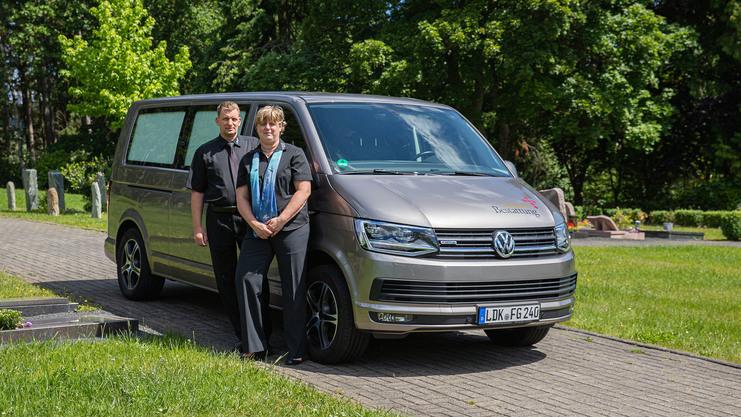 Zwei Personen in dunkler Kleidung stehen neben einem Volkswagen, umgeben von einem begrünten Friedhof.