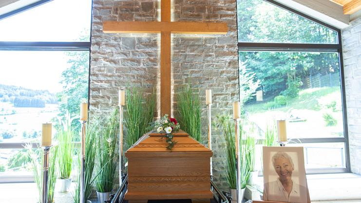 Elegante Trauerfeier mit einem geschm&uuml;ckten Holzsarg, Kerzen und einem Kreuz in einem hellen Raum mit Aussicht.