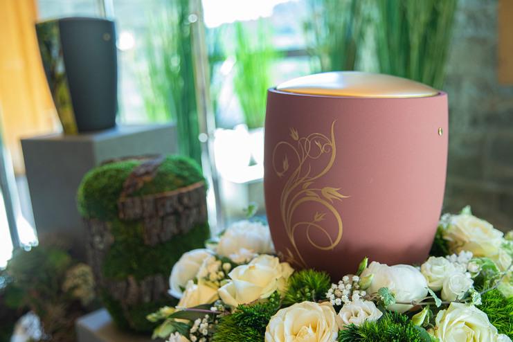 Vase mit floralen Verzierungen, umgeben von weißen Rosen und grüner Dekoration in einem hellen Raum.