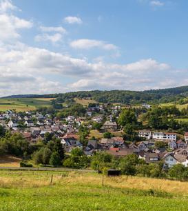 Eine Panoramafotografie von Eibach bei Dillenburg, umgeben von grünen Feldern und Wäldern unter einem bewölkten Himmel. Im Hintergrund sind Hügel und Windkraftanlagen sichtbar.
