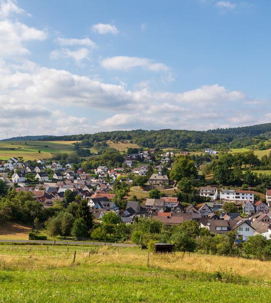 Eine Panoramafotografie von Eibach bei Dillenburg, umgeben von grünen Feldern und Wäldern unter einem bewölkten Himmel. Im Hintergrund sind Hügel und Windkraftanlagen sichtbar.