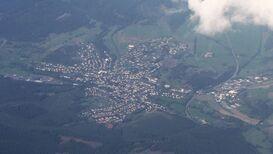 Eine Luftaufnahme des Ortes Eibelshausen, umgeben von Wäldern und Feldern. Die Siedlung ist deutlich erkennbar, mit Häusern, Straßen und einem Sportplatz. Ein Teil des Himmels ist von Wolken bedeckt.