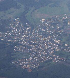 Eine Luftaufnahme des Ortes Eibelshausen, umgeben von Wäldern und Feldern. Die Siedlung ist deutlich erkennbar, mit Häusern, Straßen und einem Sportplatz. Ein Teil des Himmels ist von Wolken bedeckt.