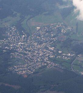 Eine Luftaufnahme des Ortes Eibelshausen, umgeben von Wäldern und Feldern. Die Siedlung ist deutlich erkennbar, mit Häusern, Straßen und einem Sportplatz. Ein Teil des Himmels ist von Wolken bedeckt.