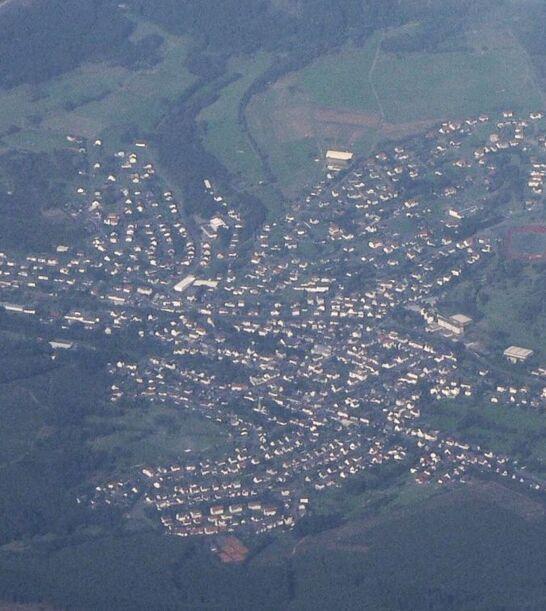 Eine Luftaufnahme des Ortes Eibelshausen, umgeben von Wäldern und Feldern. Die Siedlung ist deutlich erkennbar, mit Häusern, Straßen und einem Sportplatz. Ein Teil des Himmels ist von Wolken bedeckt.