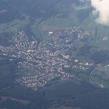 Luftaufnahme von Eibelshausen Eine Luftaufnahme des Ortes Eibelshausen, umgeben von Wäldern und Feldern. Die Siedlung ist deutlich erkennbar, mit Häusern, Straßen und einem Sportplatz. Ein Teil des Himmels ist von Wolken bedeckt.