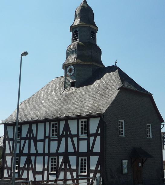 Eine Fachwerkkapelle mit Schieferdach und Turm in Mandeln. Das historische Gebäude steht an einer Straßenecke unter einem klaren blauen Himmel. Im Hintergrund sind weitere Fachwerkhäuser zu sehen.
