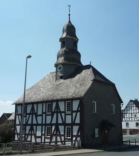 Eine Fachwerkkapelle mit Schieferdach und Turm in Mandeln. Das historische Gebäude steht an einer Straßenecke unter einem klaren blauen Himmel. Im Hintergrund sind weitere Fachwerkhäuser zu sehen.