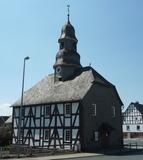 Evangelische Kapelle Mandeln Eine Fachwerkkapelle mit Schieferdach und Turm in Mandeln. Das historische Gebäude steht an einer Straßenecke unter einem klaren blauen Himmel. Im Hintergrund sind weitere Fachwerkhäuser zu sehen.