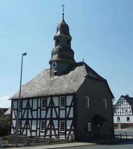 Eine Fachwerkkapelle mit Schieferdach und Turm in Mandeln. Das historische Gebäude steht an einer Straßenecke unter einem klaren blauen Himmel. Im Hintergrund sind weitere Fachwerkhäuser zu sehen.