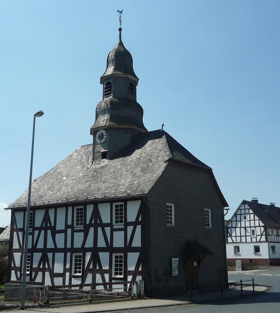 Eine Fachwerkkapelle mit Schieferdach und Turm in Mandeln. Das historische Gebäude steht an einer Straßenecke unter einem klaren blauen Himmel. Im Hintergrund sind weitere Fachwerkhäuser zu sehen.