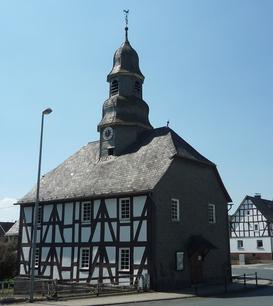 Eine Fachwerkkapelle mit Schieferdach und Turm in Mandeln. Das historische Gebäude steht an einer Straßenecke unter einem klaren blauen Himmel. Im Hintergrund sind weitere Fachwerkhäuser zu sehen.