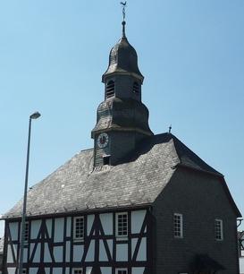 Eine Fachwerkkapelle mit Schieferdach und Turm in Mandeln. Das historische Gebäude steht an einer Straßenecke unter einem klaren blauen Himmel. Im Hintergrund sind weitere Fachwerkhäuser zu sehen.