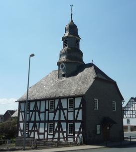 Eine Fachwerkkapelle mit Schieferdach und Turm in Mandeln. Das historische Gebäude steht an einer Straßenecke unter einem klaren blauen Himmel. Im Hintergrund sind weitere Fachwerkhäuser zu sehen.