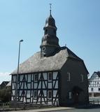 Evangelische Kapelle Mandeln Eine Fachwerkkapelle mit Schieferdach und Turm in Mandeln. Das historische Gebäude steht an einer Straßenecke unter einem klaren blauen Himmel. Im Hintergrund sind weitere Fachwerkhäuser zu sehen.