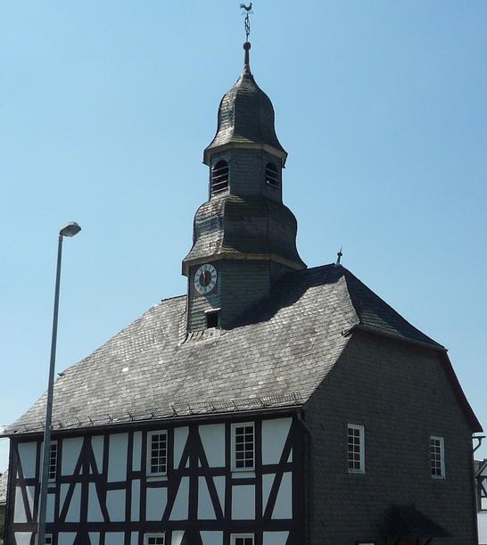Eine Fachwerkkapelle mit Schieferdach und Turm in Mandeln. Das historische Gebäude steht an einer Straßenecke unter einem klaren blauen Himmel. Im Hintergrund sind weitere Fachwerkhäuser zu sehen.