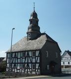 Evangelische Kapelle Mandeln Eine Fachwerkkapelle mit Schieferdach und Turm in Mandeln. Das historische Gebäude steht an einer Straßenecke unter einem klaren blauen Himmel. Im Hintergrund sind weitere Fachwerkhäuser zu sehen.