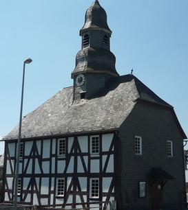 Eine Fachwerkkapelle mit Schieferdach und Turm in Mandeln. Das historische Gebäude steht an einer Straßenecke unter einem klaren blauen Himmel. Im Hintergrund sind weitere Fachwerkhäuser zu sehen.