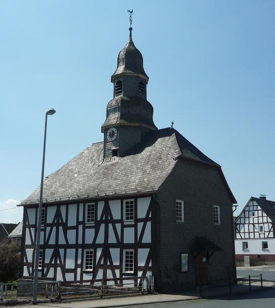 Eine Fachwerkkapelle mit Schieferdach und Turm in Mandeln. Das historische Gebäude steht an einer Straßenecke unter einem klaren blauen Himmel. Im Hintergrund sind weitere Fachwerkhäuser zu sehen.