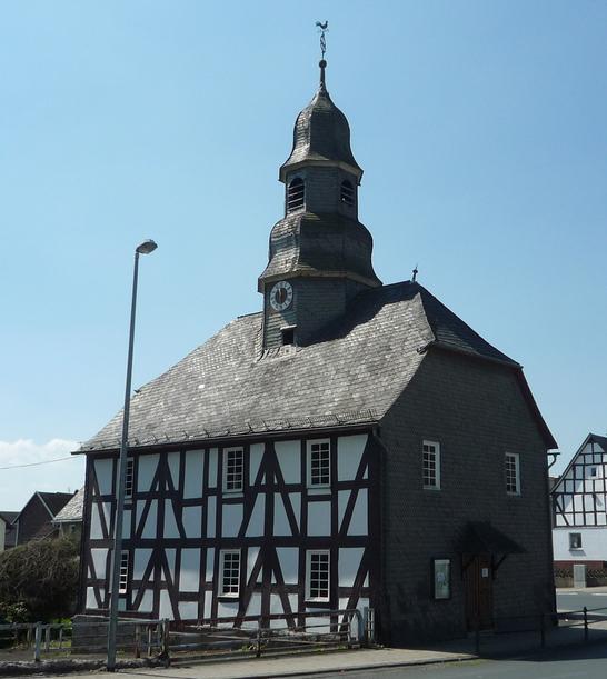 Eine Fachwerkkapelle mit Schieferdach und Turm in Mandeln. Das historische Gebäude steht an einer Straßenecke unter einem klaren blauen Himmel. Im Hintergrund sind weitere Fachwerkhäuser zu sehen.