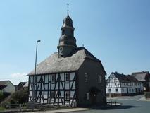Eine Fachwerkkapelle mit Schieferdach und Turm in Mandeln. Das historische Gebäude steht an einer Straßenecke unter einem klaren blauen Himmel. Im Hintergrund sind weitere Fachwerkhäuser zu sehen.