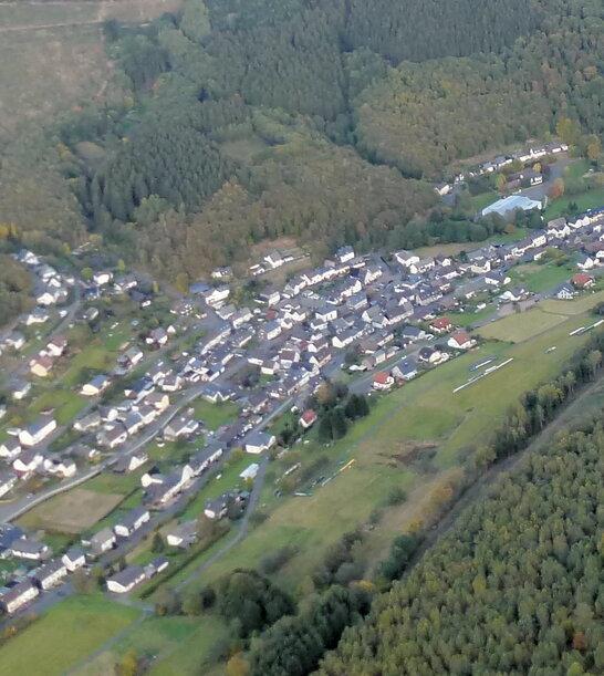 Luftaufnahme eines Dorfes umgeben von W&auml;ldern und Feldern in einer h&uuml;geligen Landschaft.