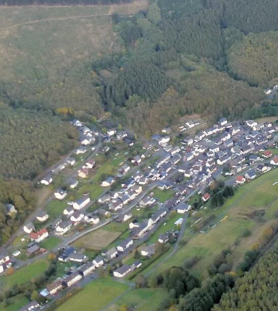 Luftaufnahme eines Dorfes umgeben von W&auml;ldern und Feldern in einer h&uuml;geligen Landschaft.