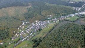 Luftaufnahme eines Dorfes umgeben von Wäldern und Feldern in einer hügeligen Landschaft.
