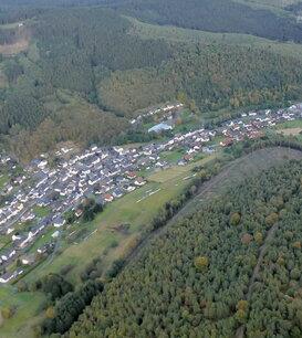 Luftaufnahme eines Dorfes umgeben von Wäldern und Feldern in einer hügeligen Landschaft.