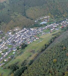 Luftaufnahme eines Dorfes umgeben von W&auml;ldern und Feldern in einer h&uuml;geligen Landschaft.