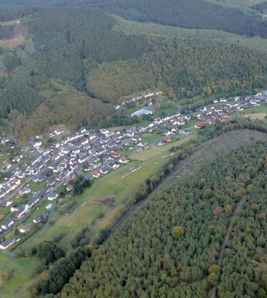 Luftaufnahme eines Dorfes umgeben von Wäldern und Feldern in einer hügeligen Landschaft.