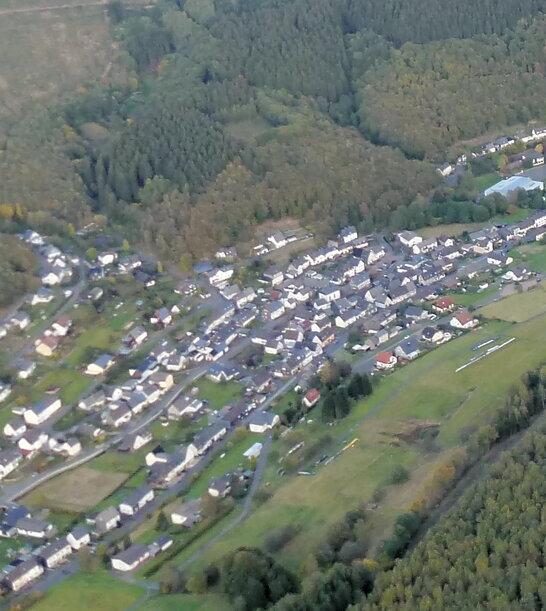 Luftaufnahme eines Dorfes umgeben von W&auml;ldern und Feldern in einer h&uuml;geligen Landschaft.