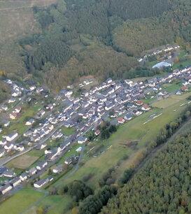 Luftaufnahme eines Dorfes umgeben von W&auml;ldern und Feldern in einer h&uuml;geligen Landschaft.