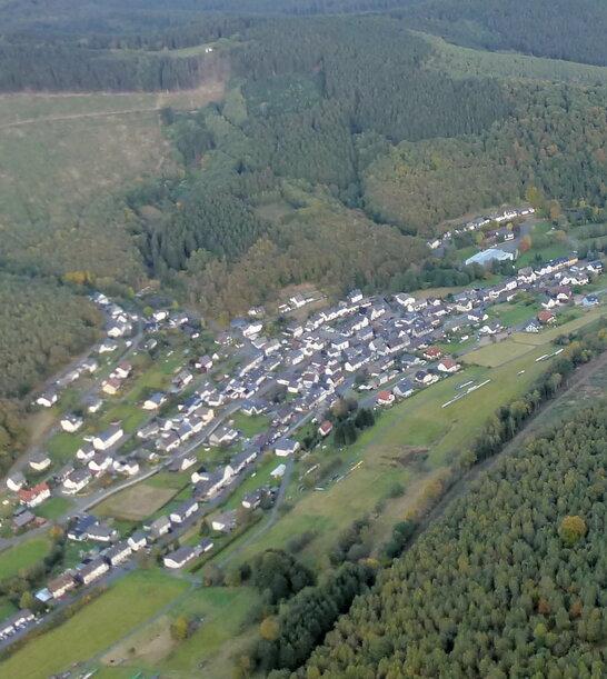Luftaufnahme eines Dorfes umgeben von Wäldern und Feldern in einer hügeligen Landschaft.