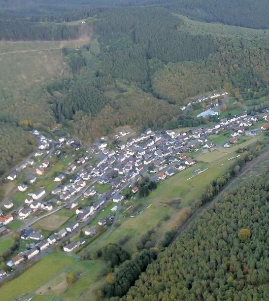 Luftaufnahme eines Dorfes umgeben von Wäldern und Feldern in einer hügeligen Landschaft.