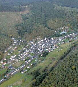 Luftaufnahme eines Dorfes umgeben von Wäldern und Feldern in einer hügeligen Landschaft.