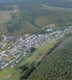 Luftaufnahme eines Dorfes umgeben von Wäldern und Feldern in einer hügeligen Landschaft.