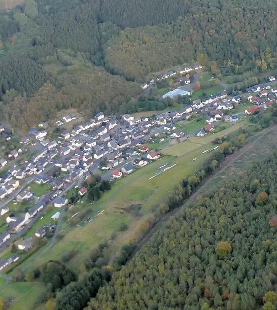 Luftaufnahme eines Dorfes umgeben von W&auml;ldern und Feldern in einer h&uuml;geligen Landschaft.