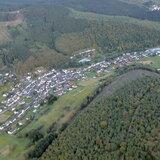 Luftaufnahme eines Dorfes umgeben von Wäldern und Feldern in einer hügeligen Landschaft.