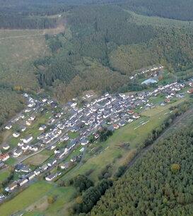 Luftaufnahme eines Dorfes umgeben von Wäldern und Feldern in einer hügeligen Landschaft.