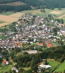 Luftaufnahme eines kleinen Dorfes umgeben von Feldern und W&auml;ldern, mit angelegten Stra&szlig;en und H&auml;usern.