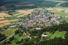 Luftaufnahme eines kleinen Dorfes umgeben von Feldern und W&auml;ldern, mit angelegten Stra&szlig;en und H&auml;usern.