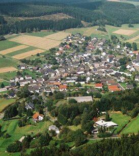 Luftaufnahme eines kleinen Dorfes umgeben von Feldern und Wäldern, mit angelegten Straßen und Häusern.
