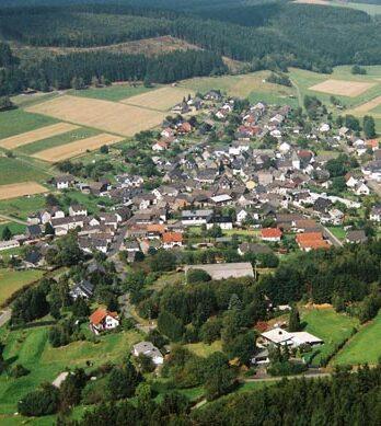 Luftaufnahme eines kleinen Dorfes umgeben von Feldern und Wäldern, mit angelegten Straßen und Häusern.