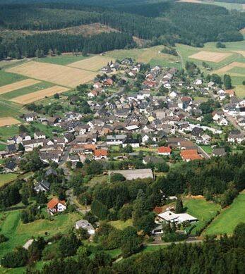 Luftaufnahme eines kleinen Dorfes umgeben von Feldern und W&auml;ldern, mit angelegten Stra&szlig;en und H&auml;usern.
